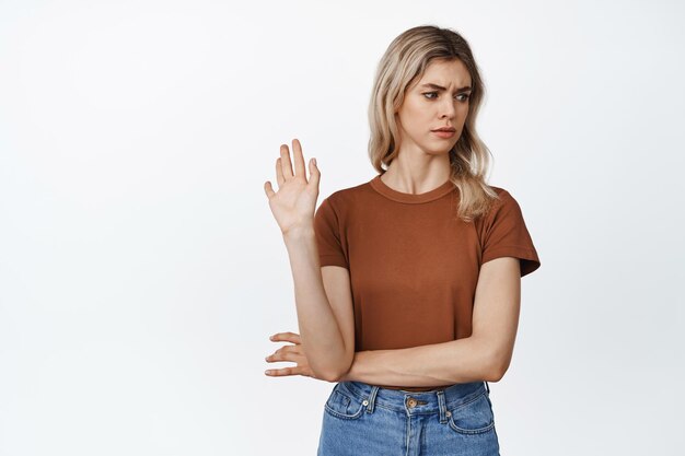 Скептически настроенная молодая женщина машет рукой, чтобы сказать, что не отказывается от чего-то с высокомерным выражением лица, недовольным предложением, стоящим на белом фоне