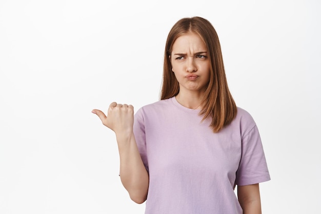 Skeptical young woman with blond hair, summer t-shirt, grimacing and furrow eyebrows from dislike, smirking unsatisfied, pointing and looking left, white background.