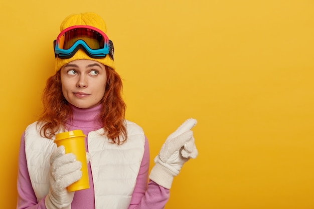 skier with ginger hair, wears yellow hat, points aside on blank space, holds takeaway coffee, demonstrates winter scenery, stands against yellow background