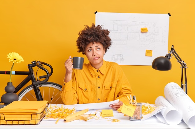 Бесплатное фото Квалифицированная профессиональная женщина-инженер пьет кофе, готовит чертежи для проекта строительства дома, глубоко задумавшись, позирует за рабочим столом с бумагами вокруг