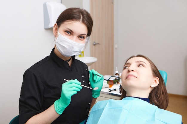 Skillful confident young female dentist wearing exam gloves and white mask holding metal probe and dental mirror, ready to examine oral cavity of woman patient sitting in chair