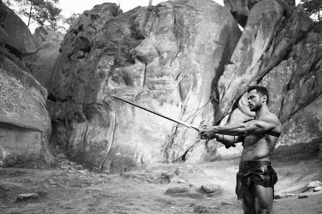 無料写真 熟練した戦闘機。岩のコピースペースの近くに立っている彼の剣を指している筋肉の強い体を持つ戦士のモノクロショット