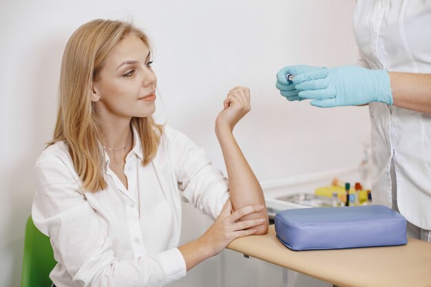 Skillful nurse is doing blood test for woman in clinic. Gilr sitting near table.