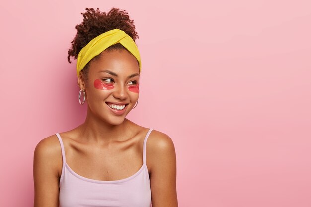 스킨 케어 개념. 눈 밑에 콜라겐 패드가있는 젊은 아프리카 계 미국인 여성은 깨끗하고 신선한 피부를 원하고 노란색 머리띠와 캐주얼 조끼를 입습니다.