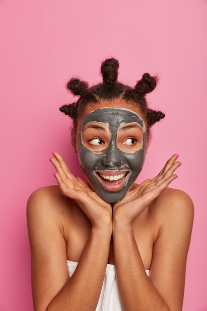 Уход за кожей, косметология и концепция благополучия. позитивная темнокожая модель кладет ладони на лицо, наносит увлажняющую маску для очищения кожи, делает косметические процедуры после ванны, накидывает на тело полотенце.