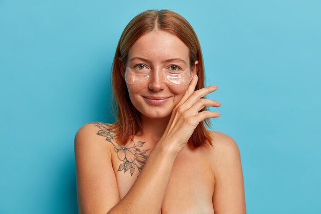 Skin care and cosmetology procedure. Satisfied freckled woman touches face gently, wears hydrogel eye patches, stands naked, has perfect well cared body with charming smile.