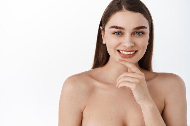 Foto gratuita cura della pelle e spa. donna di bellezza con pelle liscia e pulita, occhi azzurri e sorriso bianco perfetto, tiene la mano sul mento, sembra naturale senza trucco, spalle nude e pelle idratata e luminosa