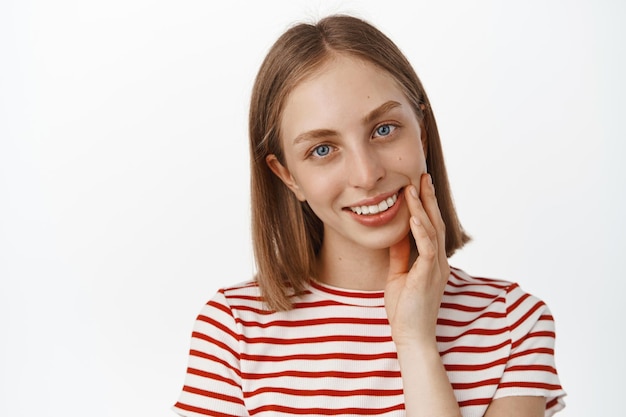 Skin care and women. Young beautiful woman with blond short hair, natural clean skin without makeup, girl touch her face cheek, smiling satisfied, standing against white background.