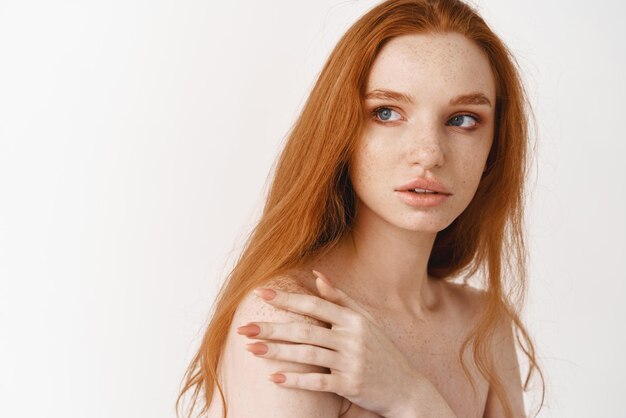 Skincare and beauty concept Closeup of tender young redhead woman with pale skin natural ginger hair touching shoulder and looking at copy space standing naked over white background