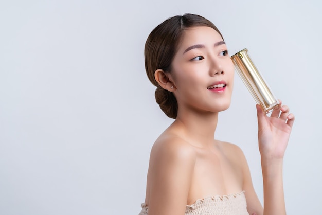 Skincare and makeup concept beautiful asian female woman with healthy facial skin close up portrait studio shot hand hold serum pr cream package with happiness and joyful