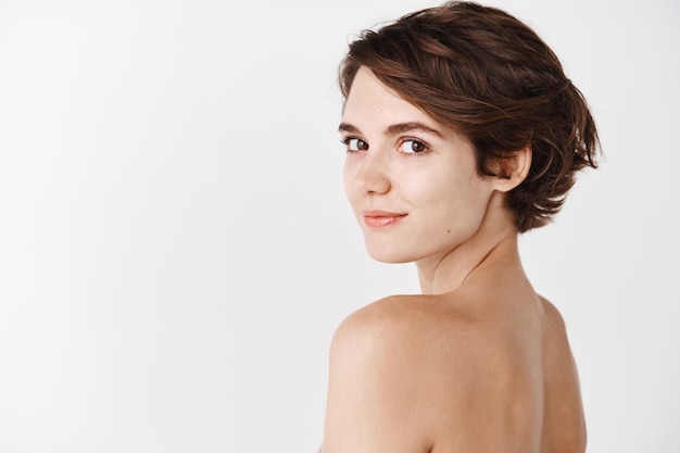 Skincare. Rear view of young caucasian woman turn head back, standing half naked on white wall and smiling. Tender girl with no makeup and natural beauty