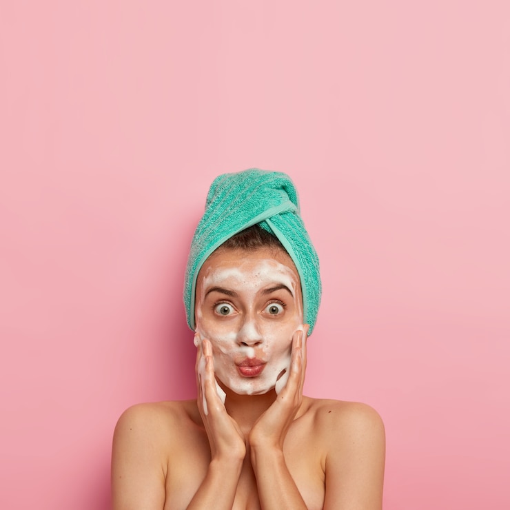 Free Photo skincare routine and hygiene concept. photo of lovely woman with widely opened eyes, keeps lips folded, massages cheeks and applies foaming soap