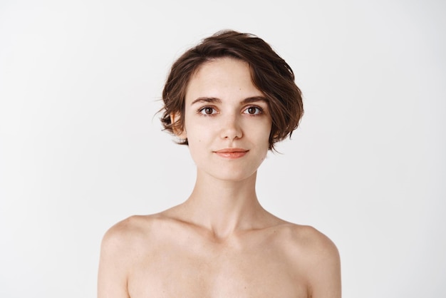 Skincare Young caucasian woman with short hair and naked body smiling at camera Girl with hydrated skin looking happy after applying daily care facial cream white background