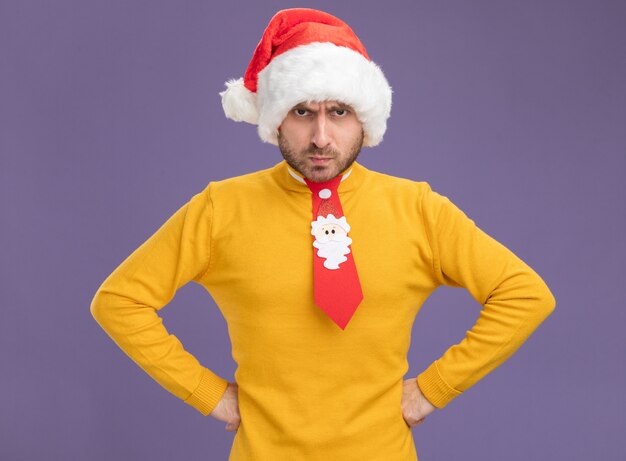 Skulking young caucasian man wearing christmas hat and tie keeping hands on waist looking at camera isolated on purple background