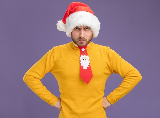 Крадущийся молодой кавказец в рождественской шляпе и галстуке держит руки на талии и смотрит в камеру, изолированную на фиолетовом фоне