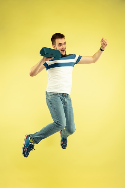 Sky sound. Full length portrait of happy jumping man with gadgets on yellow background. Modern tech, freedom of choices concept, emotions concept. Using portable speaker like superhero in flight.
