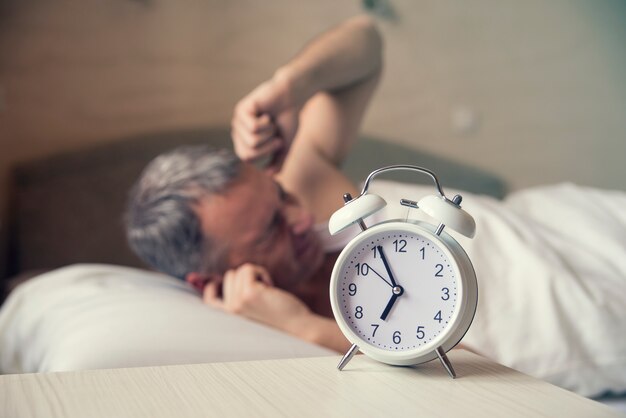 sleeping man disturbed by alarm clock early morning. Angry man in bed awoken by a noise. Waked Up. Man lying in bed turning off an alarm clock in the morning at 7am
