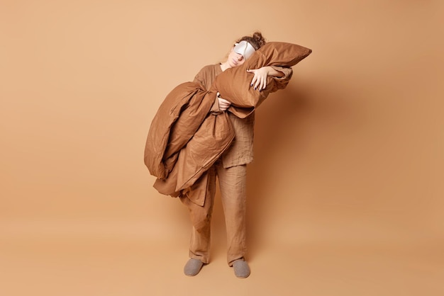 Sleeping routine and wellbeing concept Relaxed young woman leans on soft pillow wears pajama and slippers blindfold on eyes takes nap isolated over beige background enjoys peaceful atmosphere