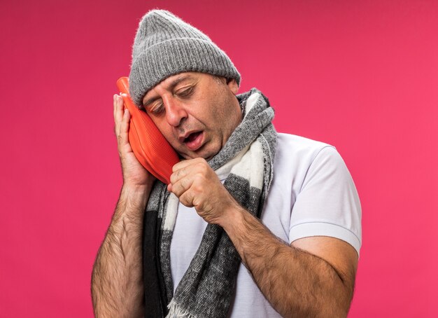 sleepy adult ill caucasian man with scarf around neck wearing winter hat coughing and holding hot water bottle isolated on pink wall with copy space