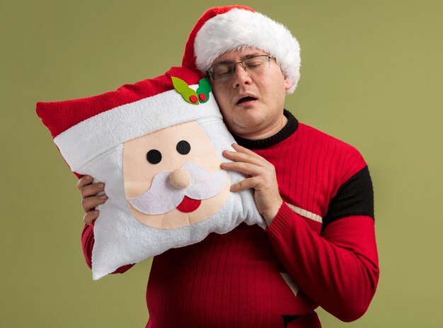 sleepy adult man wearing glasses and santa hat holding santa claus pillow touching head with it sleeping isolated on olive green background