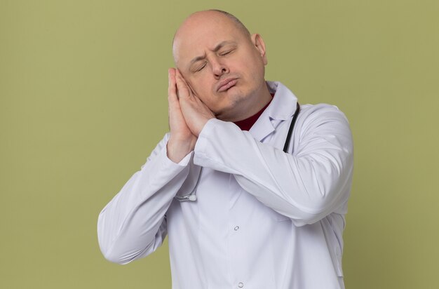 Sleepy adult slavic man in doctor uniform with stethoscope putting head on his hands 