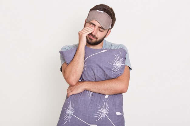 Sleepy bearded male posing with closed eyes isolated over white wall, rubbing his eyes with hand, embracing pillow