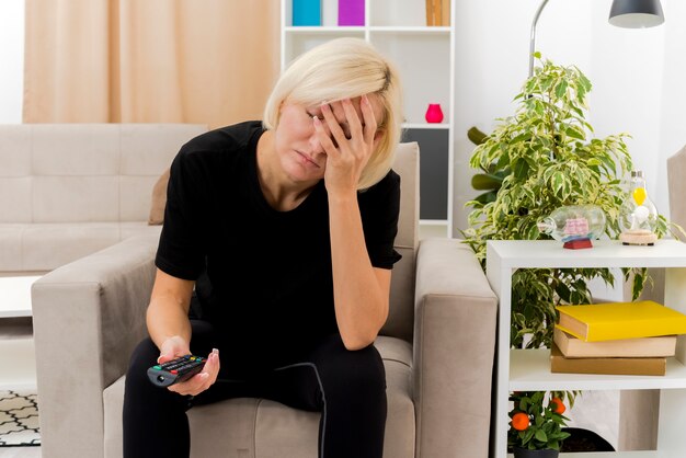 Sleepy beautiful blonde russian woman sits on armchair with closed eyes putting hand on face holding tv remote 