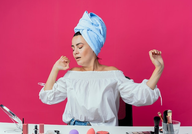 Free photo sleepy beautiful girl sits at table with makeup tools yawns isolated on pink wall