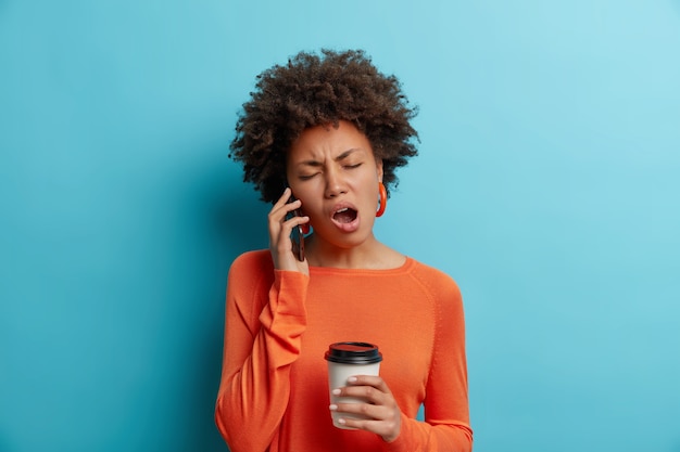 Sleepy bored Afro American woman listens uninteresting story while talking via smartphone drinks takeaway coffee wears orange jumper with earrings isolated over blue wall