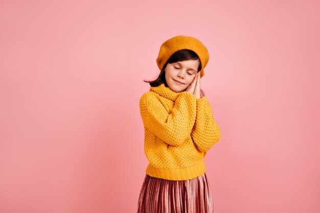 Sleepy brown-haired child standing on pink wall.  kid posing with eyes closed.