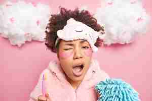 Foto gratuita la donna afroamericana riccia sonnolenta si sottopone alle procedure di igiene quotidiana al mattino tiene lo spazzolino da denti e la spugna da bagno sbadiglia con la bocca spalancata si sveglia presto al mattino pose contro il muro rosa