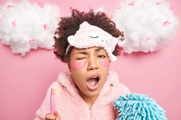 Sleepy curly Afro American woman undergoes daily hygiene procedures in morning holds toothbrush and bath sponge yawns with wide opened mouth awakes early in morning poses against pink wall