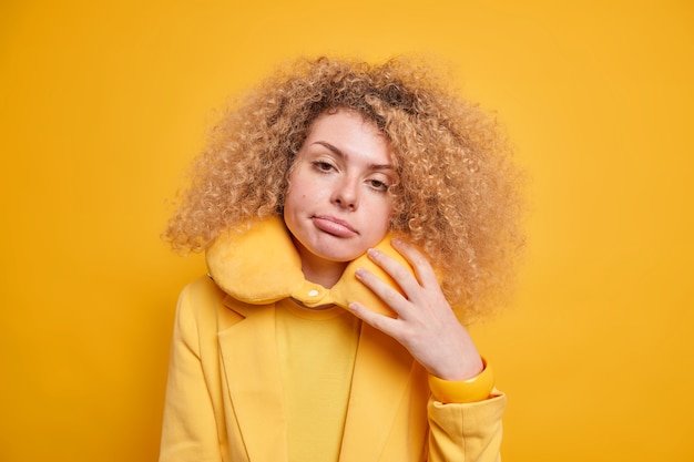 Sleepy curly haired woman looks with tired expression feels exhausted after long trip wears inflated pillow around neck for comfort tilts head dressed formally isolated over yellow wall