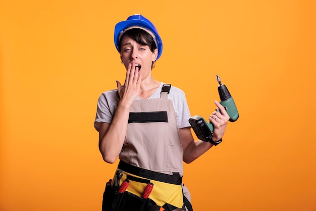 Free photo sleepy female construction worker yawning and being tired while she works on renovating or building. falling asleep and feeling exhausted, holding power drilling nail gun in studio.