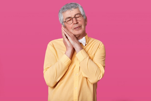 Sleepy grey haired mature man wears yellow shirt with bow tie poses with hands together while standing with closed eyes on rose wall. Male with short hairstyle wants to go to bad. People concept.