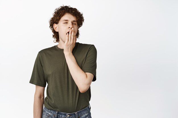 Sleepy male student with curly hair yawning with tired face yawn from boredom standing in tshirt over white background