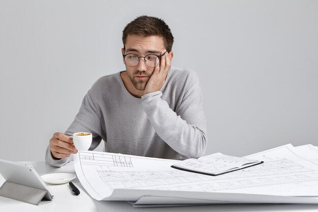 Sleepy man drinks coffee as feels tired, works all day at blueprints, has fatigue expression