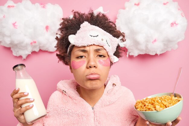 Sleepy sulking Afro American woman with sad expression wears nightwear eats healthy food applies collagen patches under eyes poses against pink wall