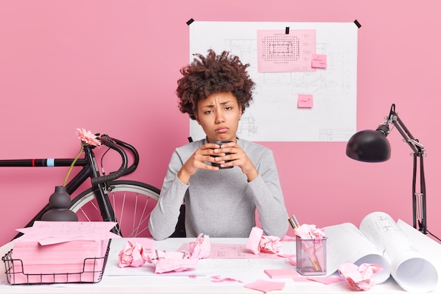 Sleepy tired woman drinks coffee poses at desktop with paper scraps works all day long on making construction project prepares architectural sketches sits in coworking space has creative skills