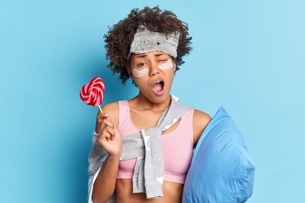 Sleepy tired woman yawns poses late at night with delicious candy holds pillow undergoes beauty procedures dressed in pajama isolated over blue wall