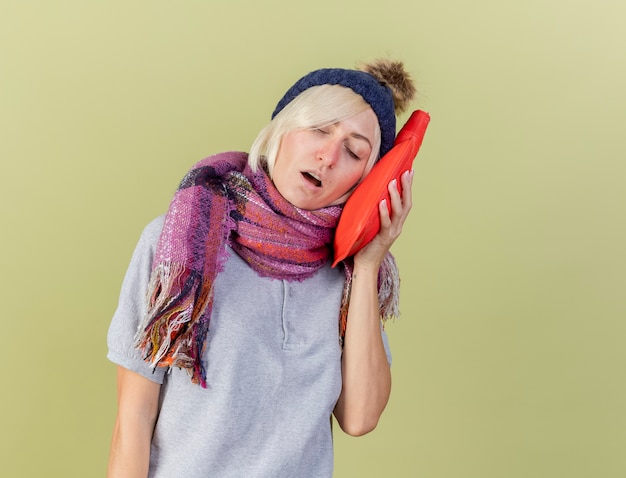 Бесплатное фото Сонная молодая блондинка больная славянская женщина в зимней шапке и шарфе кладет грелку на лицо, изолированное на оливково-зеленой стене с копией пространства