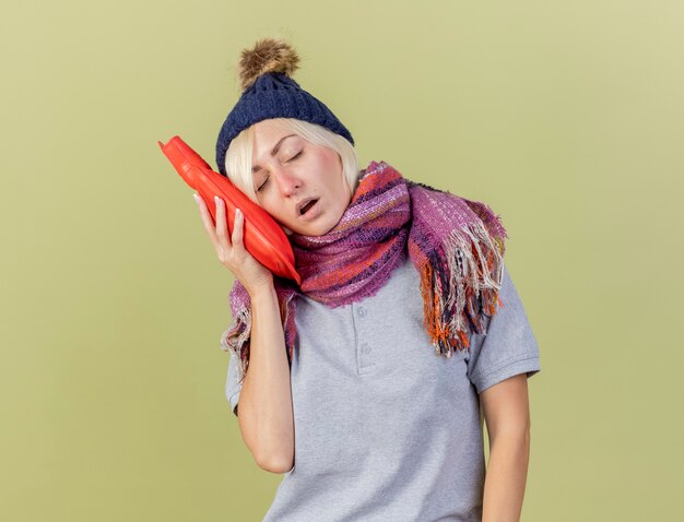 Sleepy young blonde ill slavic woman wearing winter hat and scarf stands with closed eyes putting head on hot water bottle isolated on olive green wall with copy space