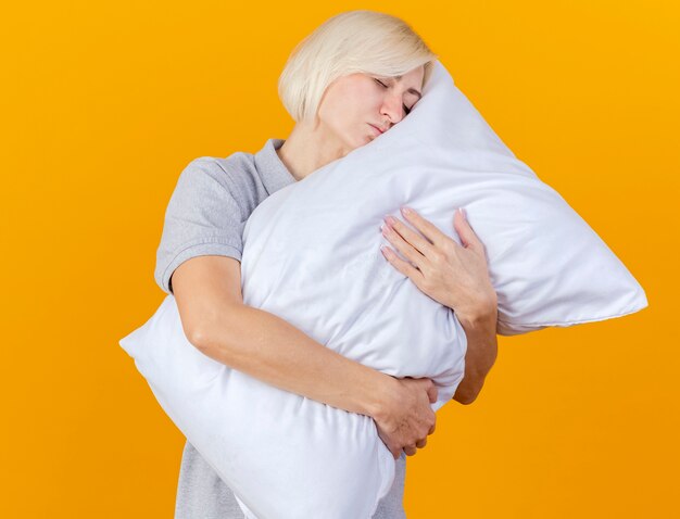 Sleepy young blonde ill woman hugs and puts head on pillow isolated on orange wall