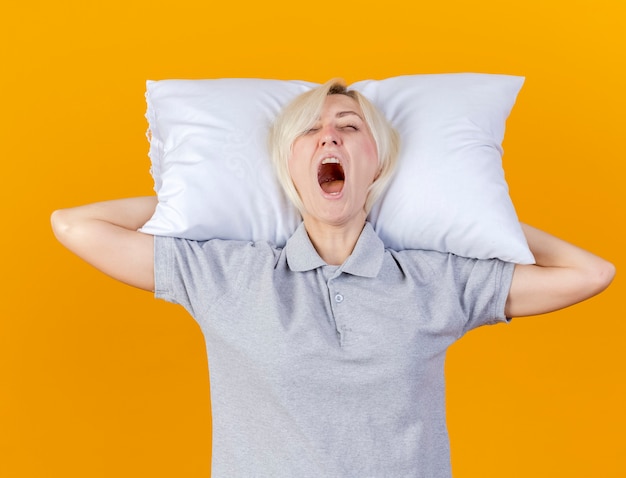 眠そうな若い金髪の病気の女性はあくびをして、オレンジ色の壁に隔離された頭の後ろに枕を保持します