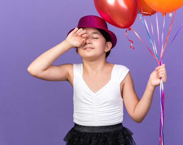 sleepy young caucasian girl with violet party hat holding helium balloons isolated on purple wall with copy space