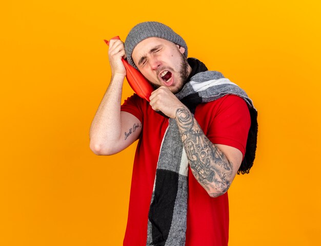 Sleepy young caucasian ill man wearing winter hat and scarf yawns and puts head on hot water bottle isolated on orange wall with copy space