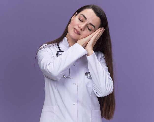 Sleepy young female doctor wearing medical robe with stethoscope puts head on hands isolated on purple wall with copy space