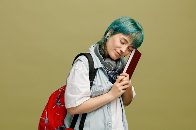 Sleepy young female student wearing headphones and bandana on neck and backpack standing in profile view holding note book putting head on it with closed eyes isolated on olive green background