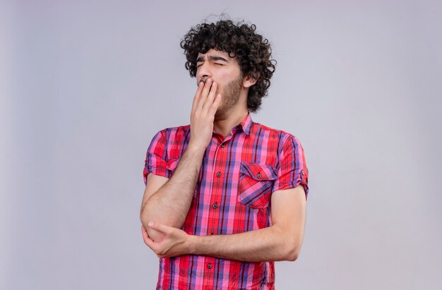 A sleepy young good-looking man with curly hair in checked shirt holding hand on mouth yawning 