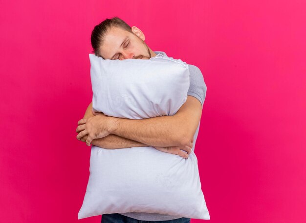 Sleepy young handsome slavic ill man hugging pillow putting head on it with closed eyes isolated on pink wall with copy space